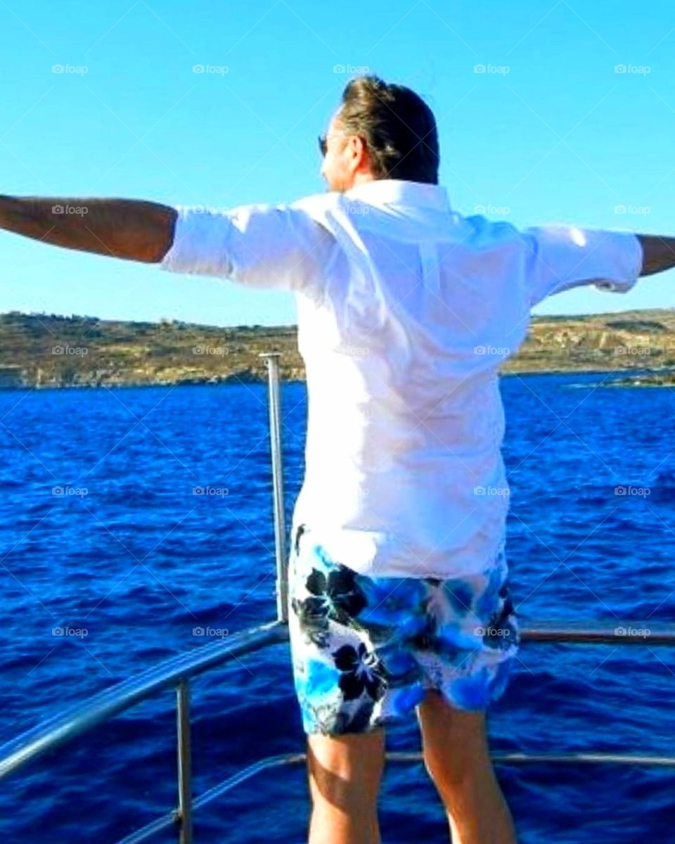 Summer.  Sea.  At the bow of the boat is a man in a white shirt.  The man spread his arms towards the sun and the wind