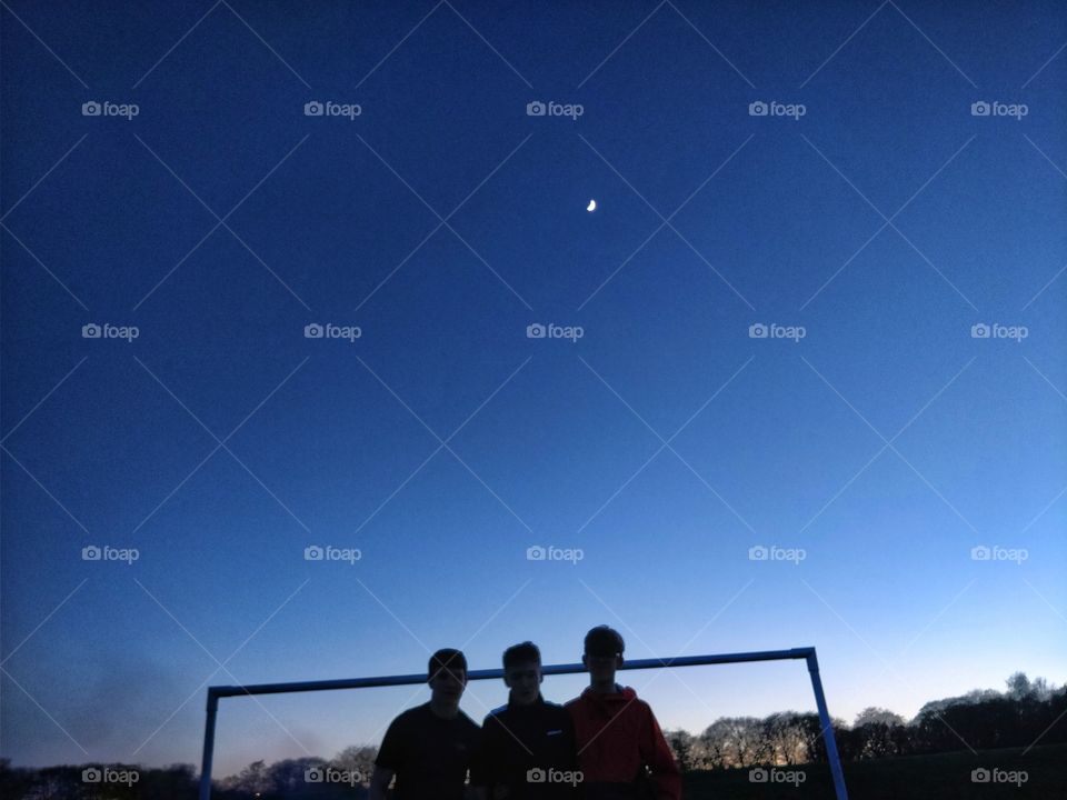 silhouetted figures, clear sky, moonlit, net on a football pitch, bit of smoke from a BBQ