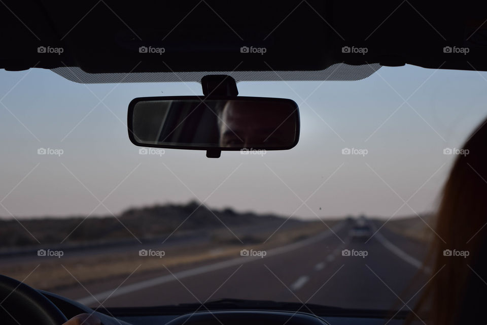 On the road. Através do parabrisa, vemos a rodovia e o céu anoitecendo e no espelho do carro, vemos o reflexo dos olhos de um homem dirigindo.