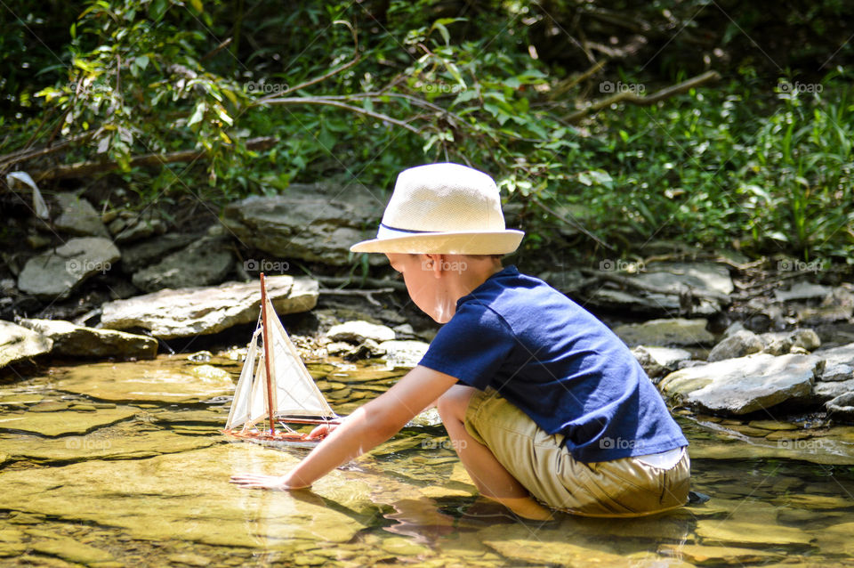 Young boy playing with a wooden sailboat in a creek on a sunny day
