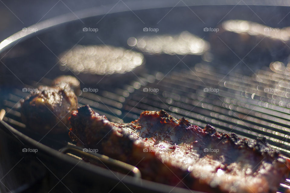 A portrait of some meat lying on a barbecue grill. there are a few different kinds. chicken drums, spare ribs and hamburgers. it brings back the summertime.