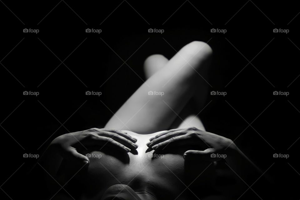 Black and white view of naked woman lying with hands on breasts on black background.