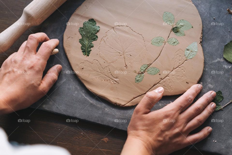 Young attractive woman in white shirt ceramic artist decorating clay plate with dry leaves at the table in pottery workshop. Handmade work student