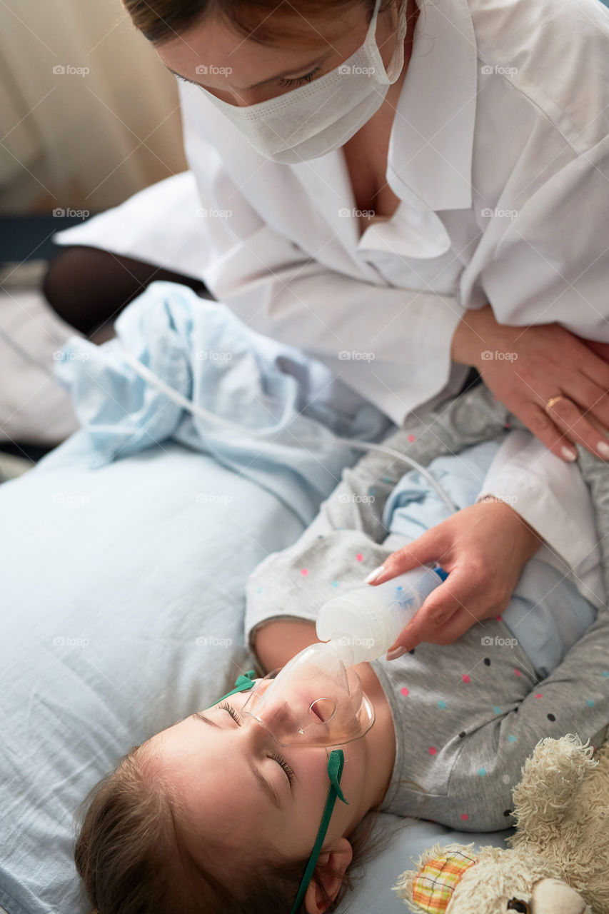 Doctor visiting little patient at home. Child having medical inhalation treatment with nebuliser. Woman wearing uniform and face mask