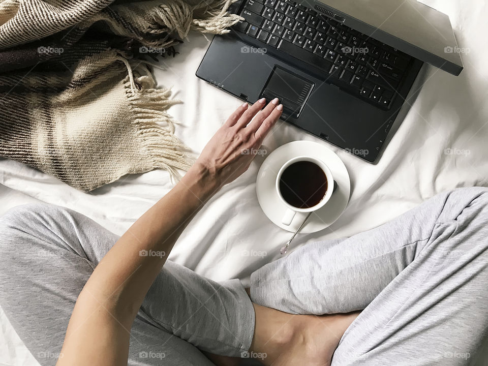 Overheard view of a young woman using a laptop and drinking morning coffee in cozy bed 