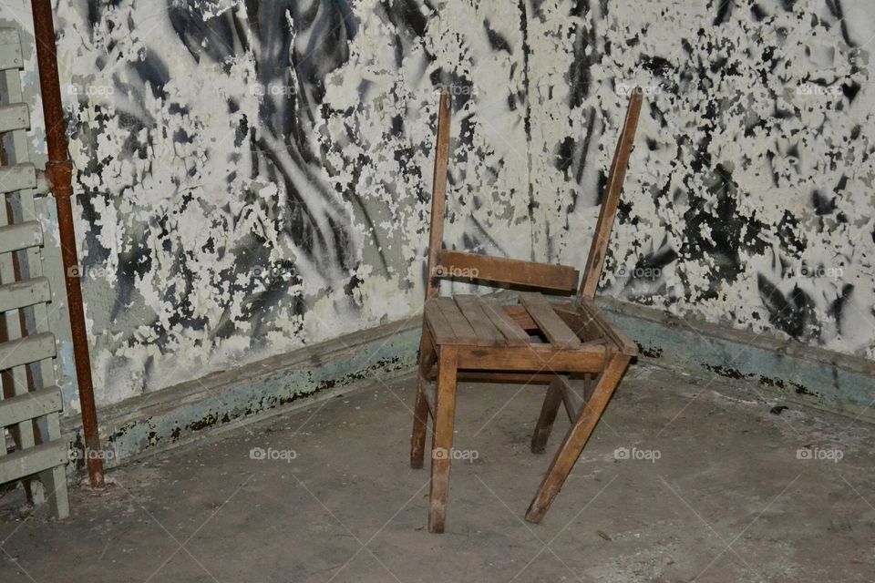 A small broken wooden chair leaning sideways in an empty room with peeling paint and a dirty floor