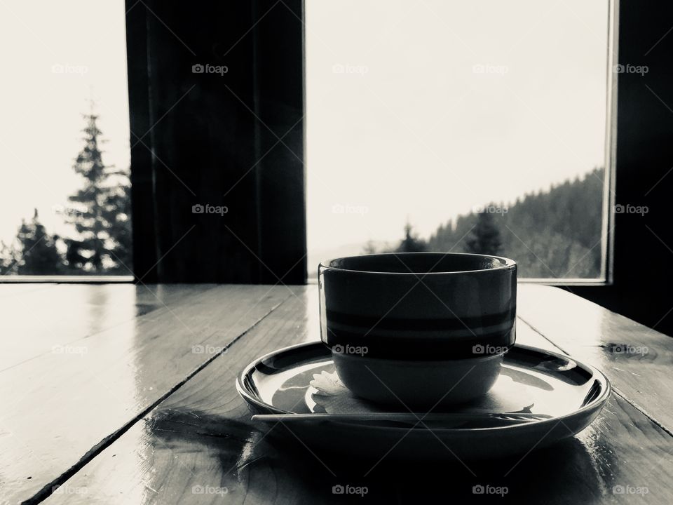 Black and white photo of cup of coffee with plate placed on wooden table near the window