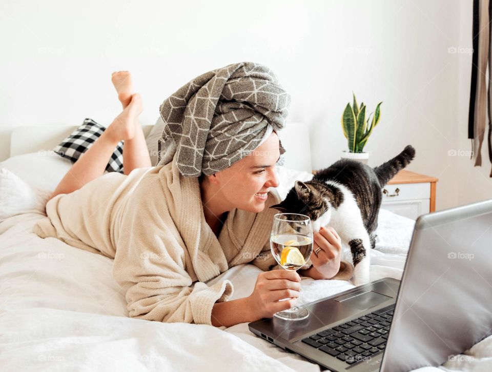 Happy young woman wearing a bath robe laying in bed while using laptop and petting her cat