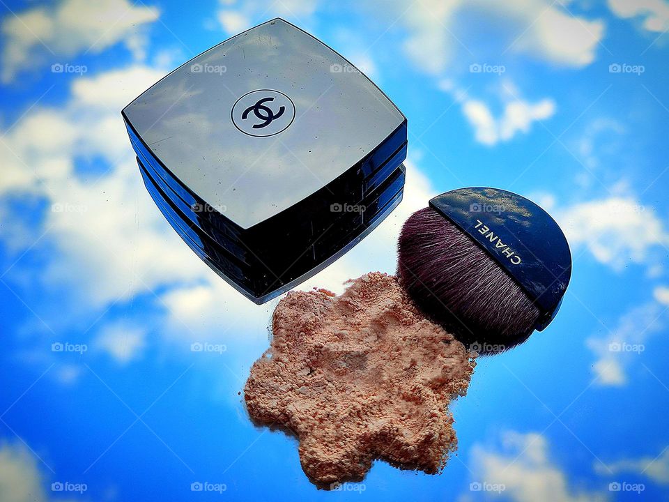 On the mirror surface is a box with the Chanel logo, a brush with the inscription "Chanel" and powder in the form of a flower against the background of the reflection of the blue sky with white clouds. Powder Chanel Les Beiges