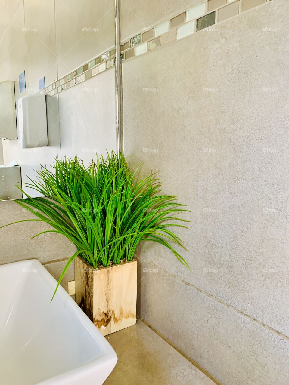 A green synthetic plant in a wooden pot in a corner of a bathroom, next to a white hand washing basin and a big mirror. 