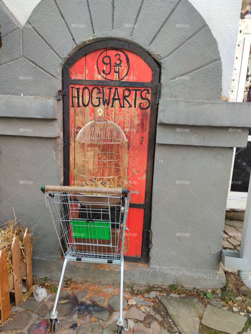 Red wooden door with black "HOGWARTS" lettering. Attached to the door is a shopping cart containing old suitcases and a metal bird cage