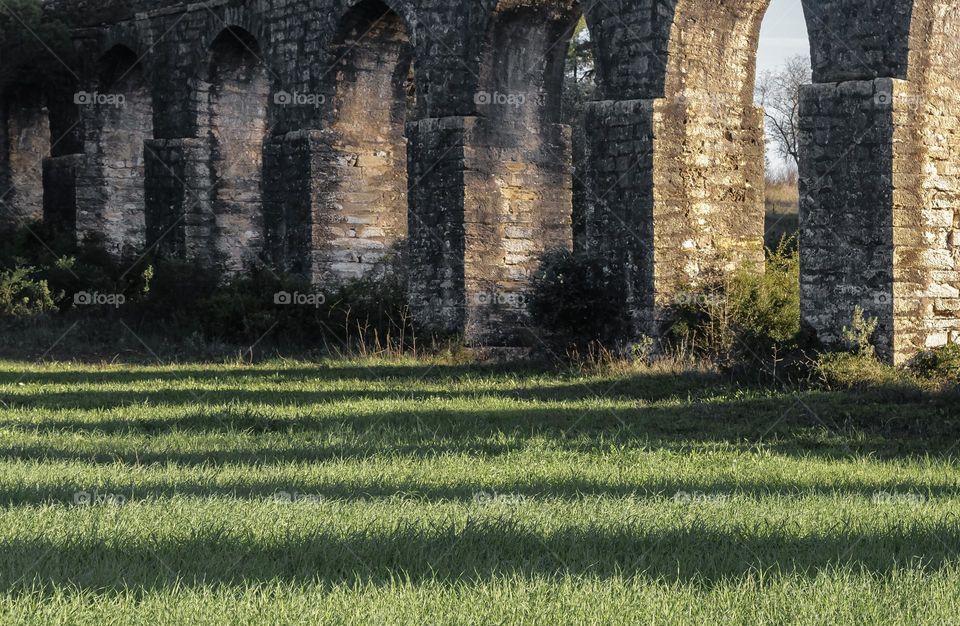 Late afternoon sun spills through the arches of an aqueduct creating shadows on the green grass