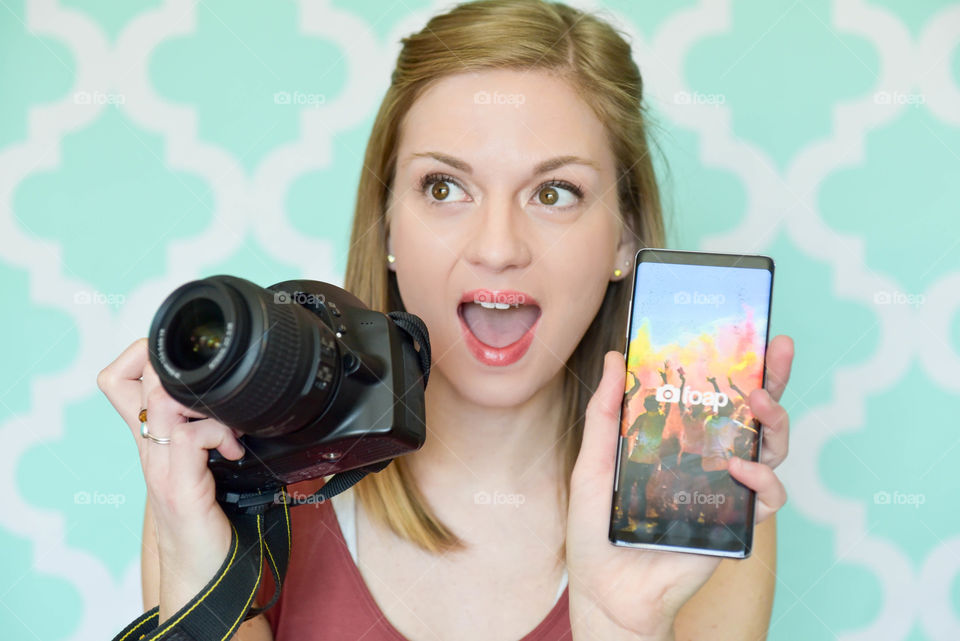Young woman happily holding up her phone with the foap app displayed in one hand and holding her DSLR camera in the other