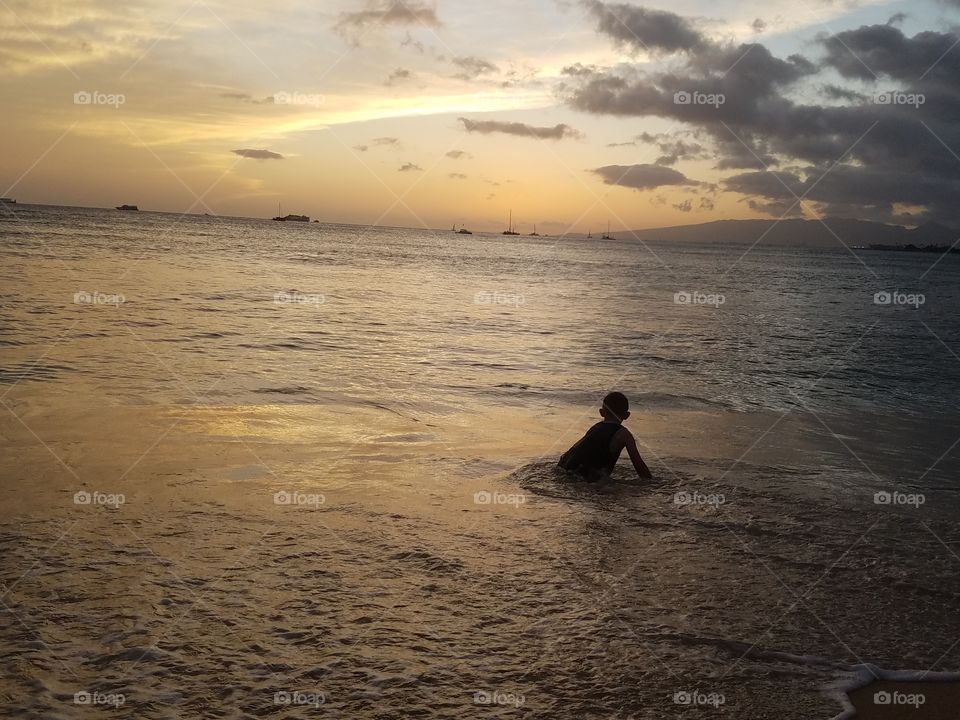 Boy sits in ocean near seashore as sun sets