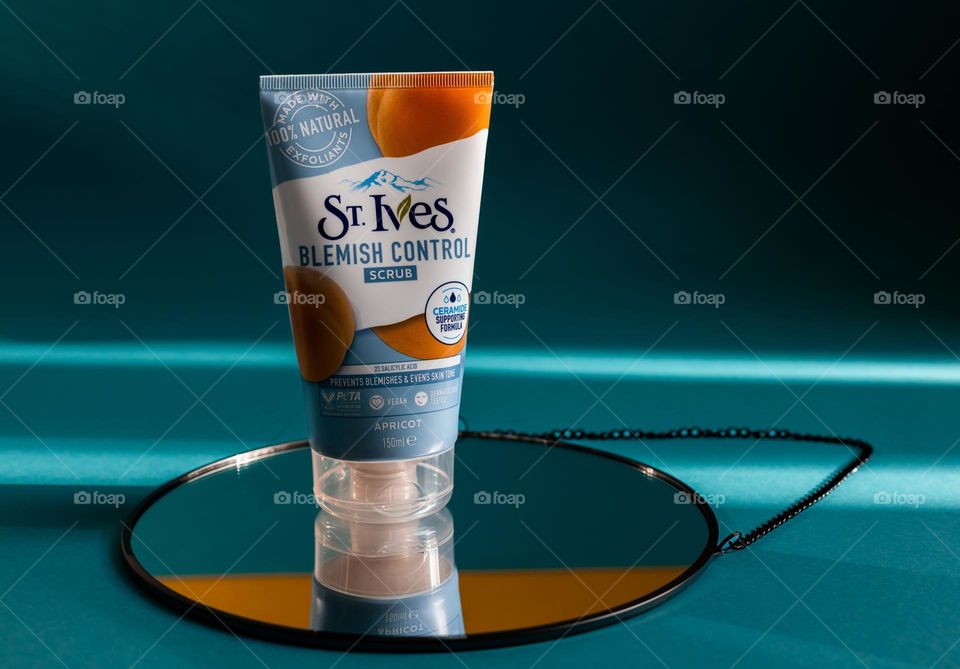 One tube of natural apricot scrub brand st. Ives stands on a round mirror with a black border and chain on a blue background with shadows and light from the sun, close-up side view. Female cosmetics concept, body care, beauty.