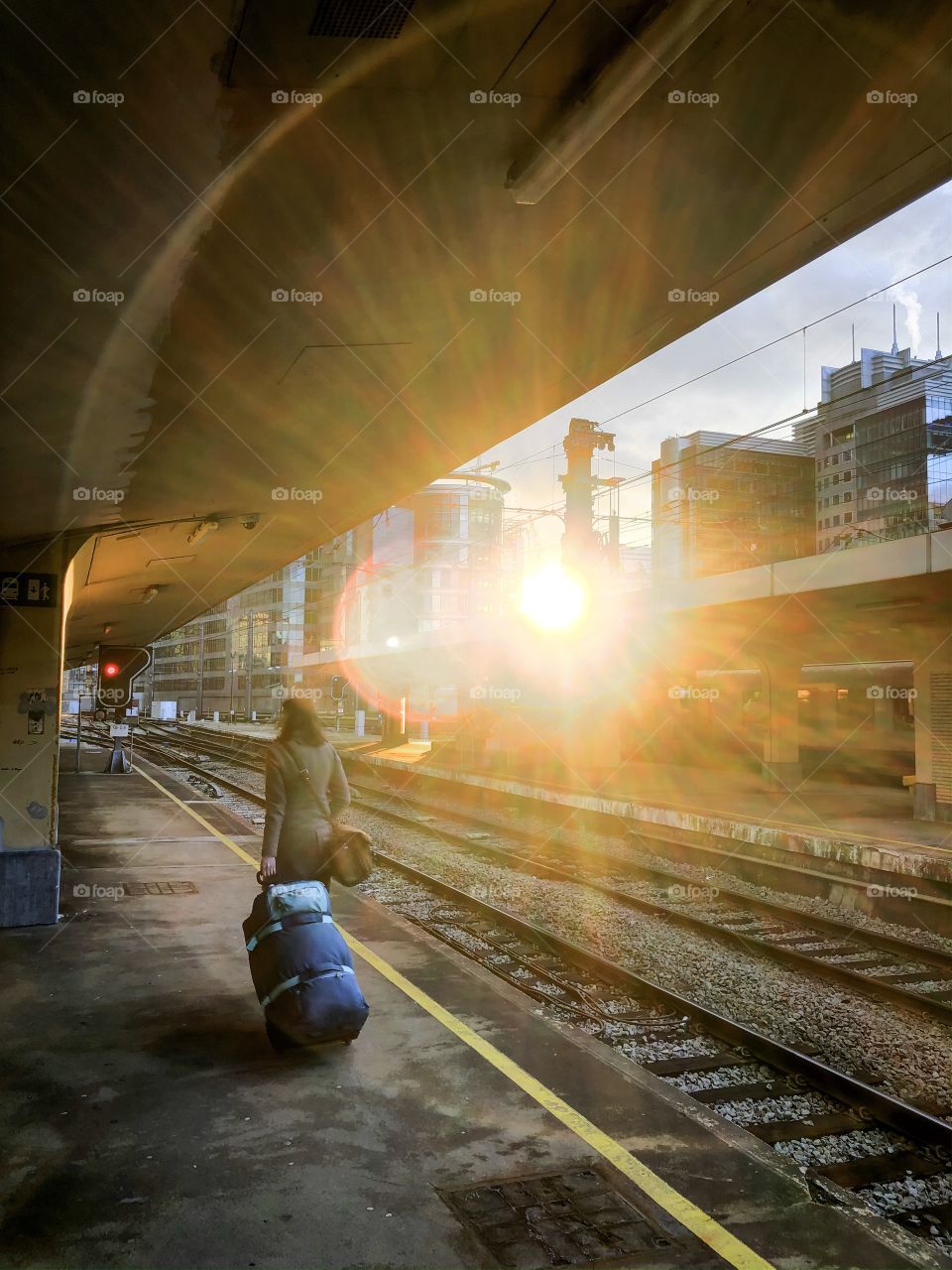 An unrecognizable young woman carrying her luggage over the platform of a railway station covered in an bright and colorful halo of light from the setting sun