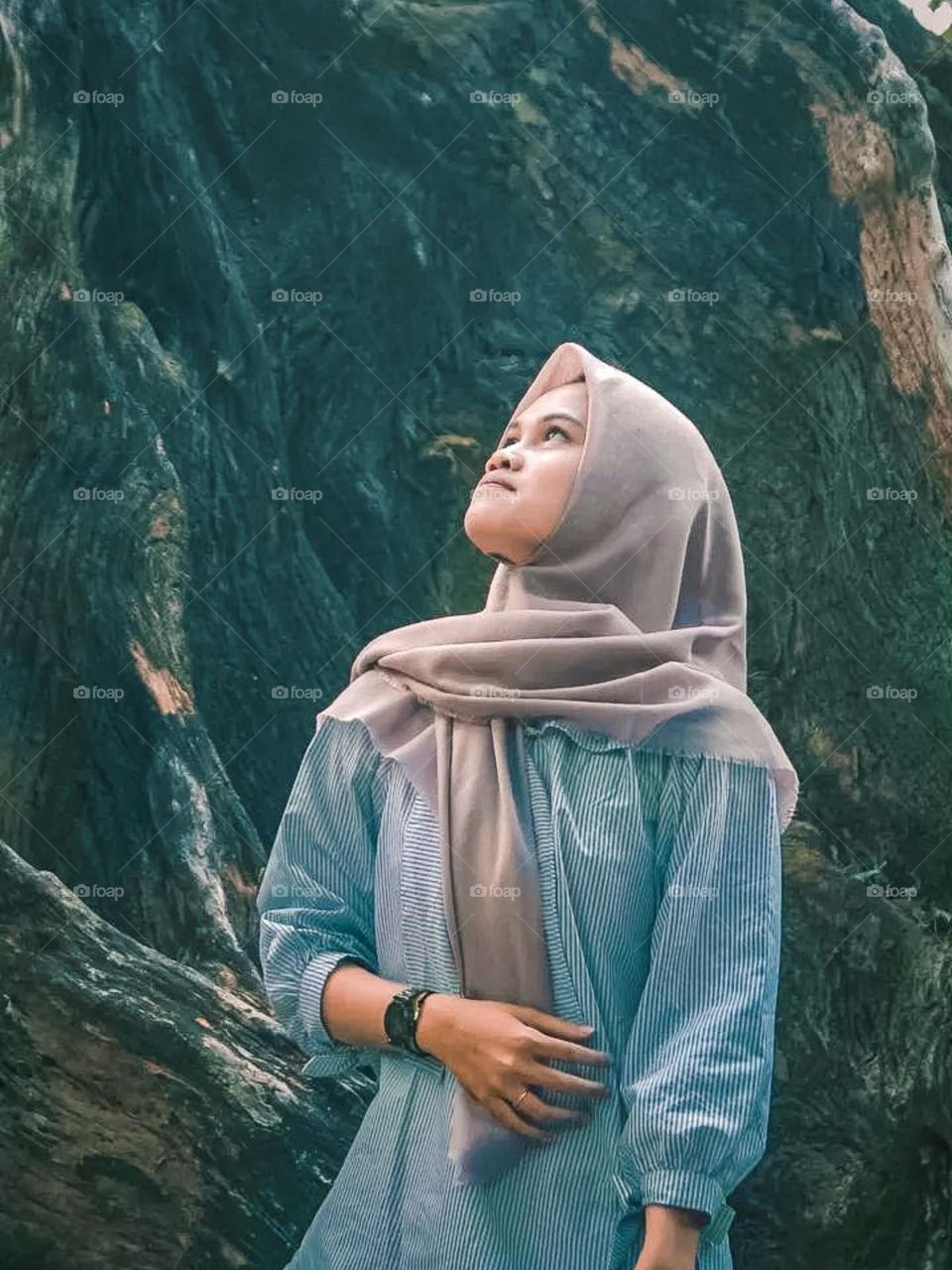 Portrait of young woman wearing hijab and striped shirt, standing in front of a background that looks like a rock or cliff