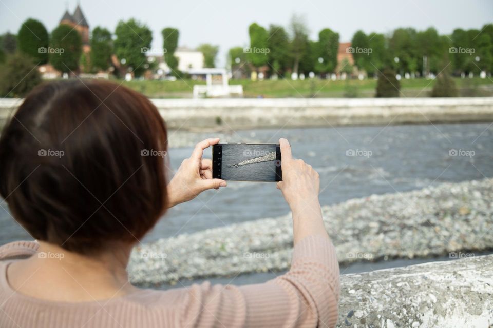 woman taking a video with a mobile phone camera, standing on the river bank at sunny spring day . Video creator concept.