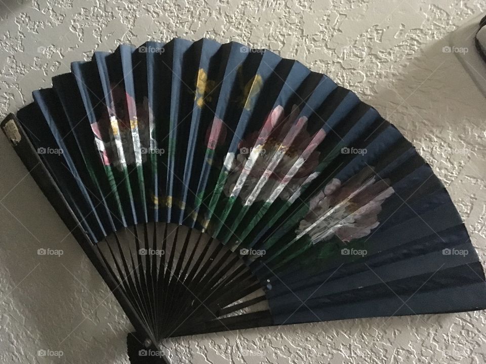 Oriental style wall fan accenting a textured white wall. Color dark green with oriental flowers.