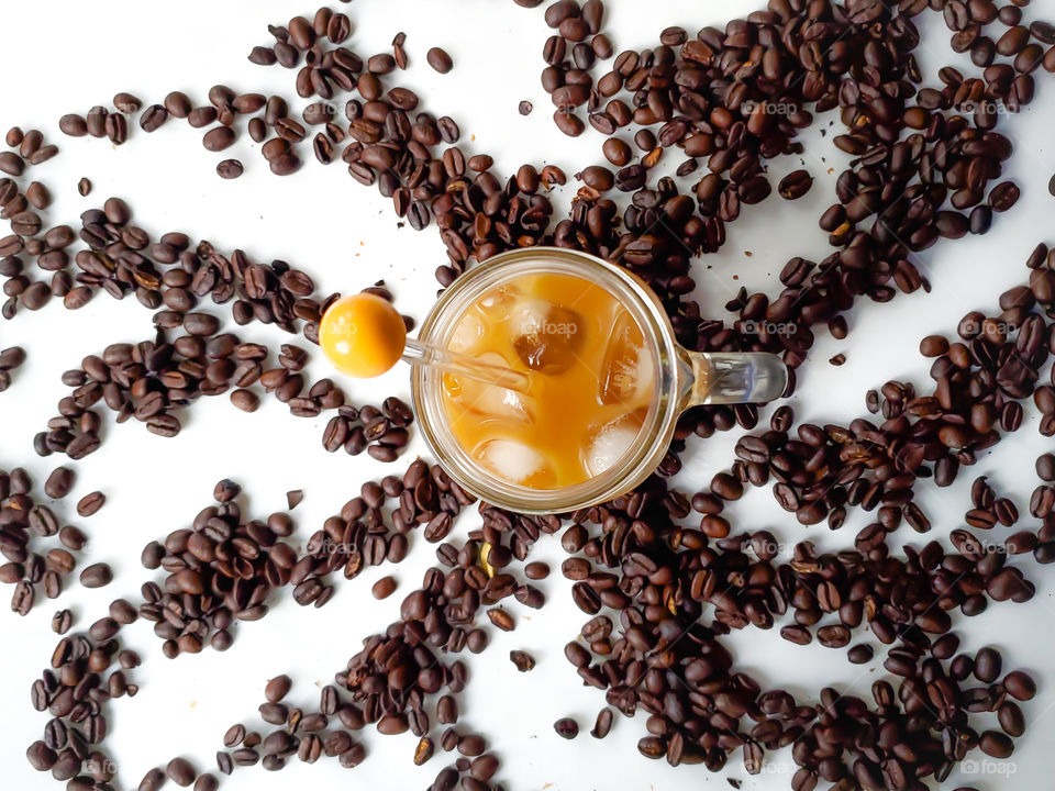 Flatlay of one iced coffee in a mason jar surrounded by a swirl of whole coffee beans on a white background.