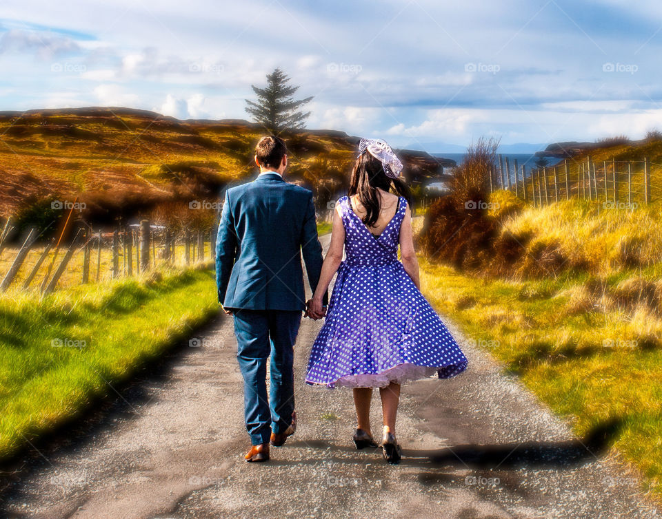A newlywed husband and wife walk away from the camera, hand in hand on the same path together, surrounded by picturesque countryside 