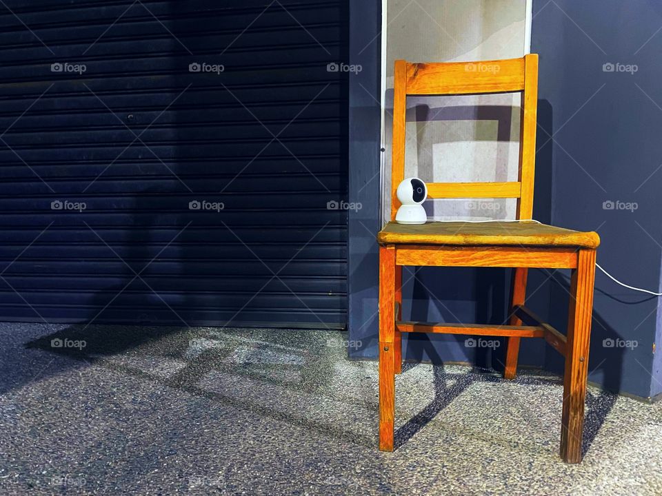 A CCTV camera perched precariously on a wooden chair. The CCTV camera faces out into the street, while the store behind it is closed.