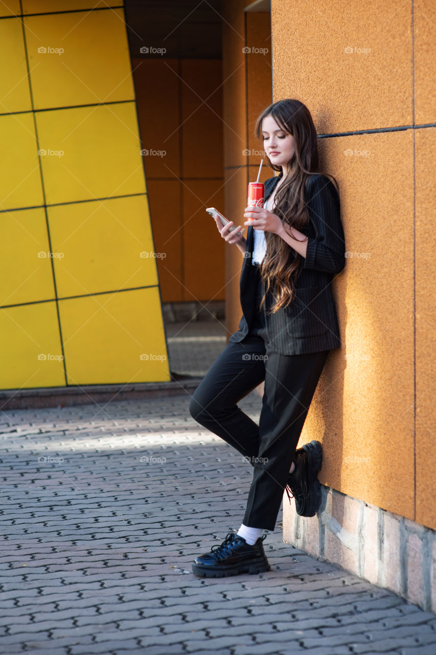 Dark hair girl drinks coca cola. Long brown hair. Teenager. Young fashion style. Using mobile phone