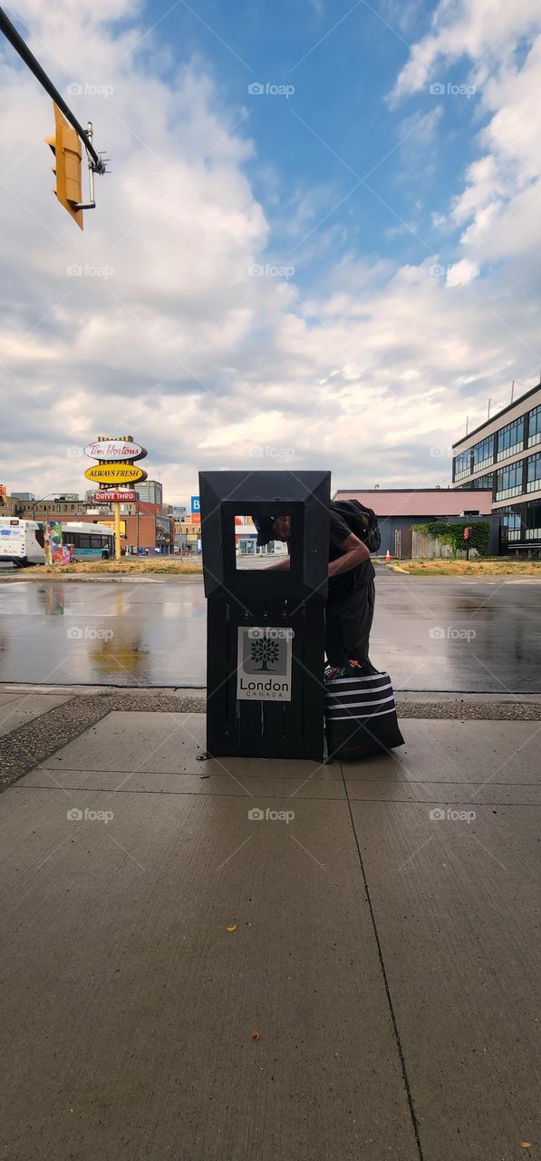 a man is in the street opening up a public garbage can and investigating its contents. it has just rained and there is a dramatic sky. the man is filling up his black reusable Store bag with items from the garbage.