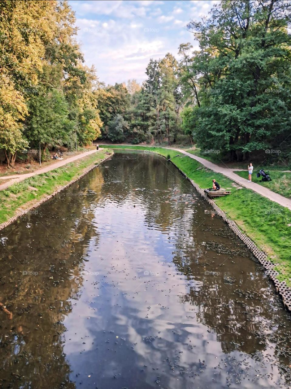 A serene park scene with a calm river flowing through lush greenery, featuring pathways on either side, visitors relaxing, and a peaceful ambiance under a bright sky.