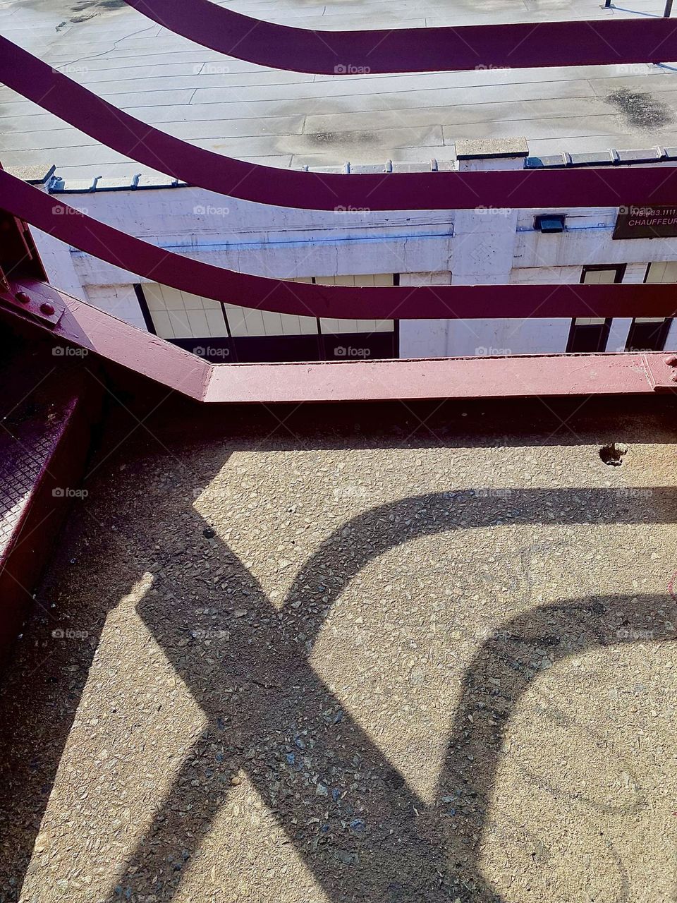 This interesting streamlined design emerges when the red metal staircase of the „Pulaski Bridge“ at „Newtown Creek“ in LIC, in particular the railing with its parallel bars casts shadows onto the paved over platforms. 2023. Hypnotic Productions
