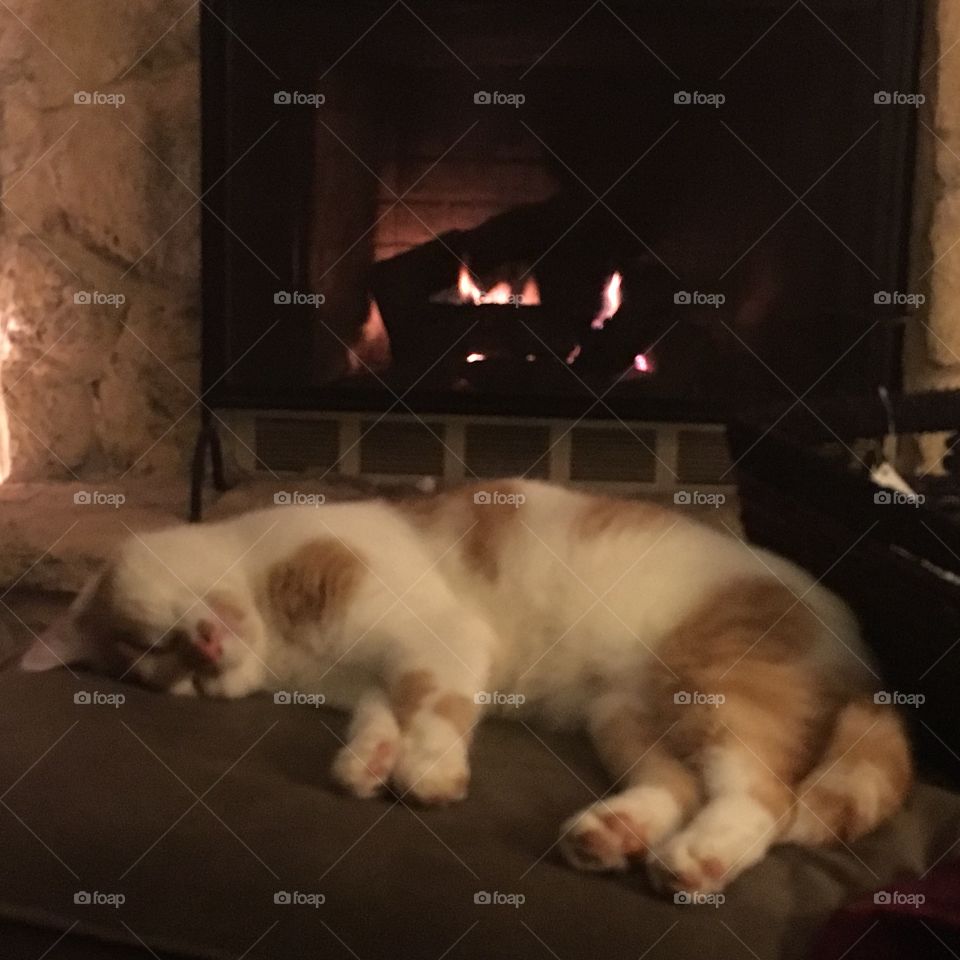 An orange and white cat sleeps on an ottoman in front of a fireplace.