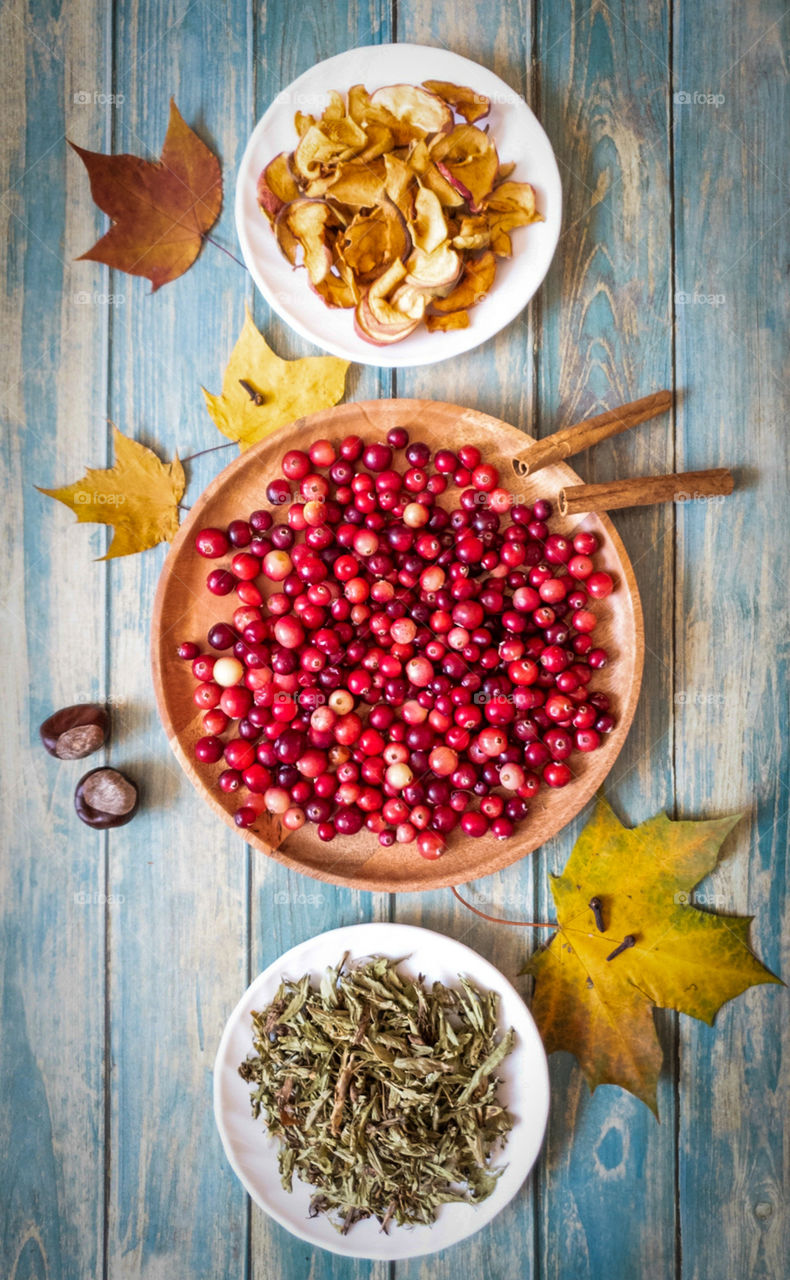 Ingredients for making healthy compote with autumn berries without added sugar. A wooden dish with cranberries, a saucer with stevia, a saucer with dried apples, cinnamon sticks on a wooden background with dry leaves and chestnuts