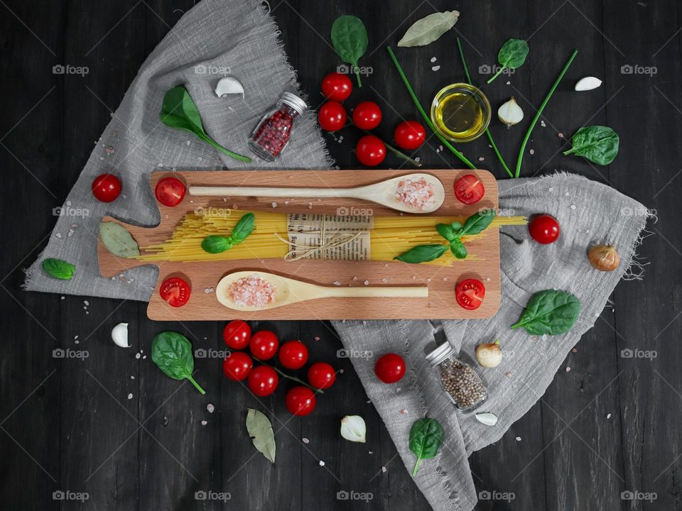 Raw spaghetti pasta on a cutting board with wooden spoons of salt, cherry tomatoes, greens, garlic, spices, leaves, basil and olive oil lie on a black wooden background, flat lay close-up.