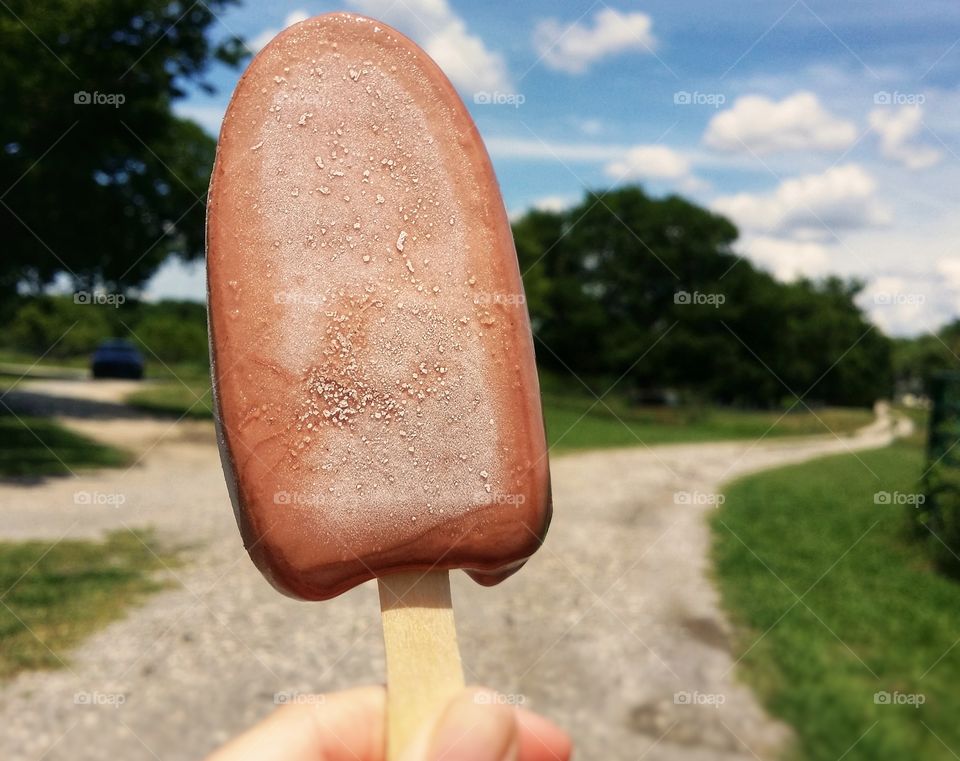 Fudge ice cream bar melting and frosty in the summer sun down a country road with green grass and a blue sky with cloudy sky