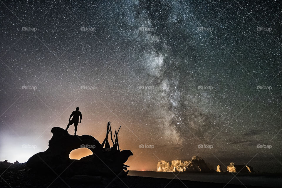 Silhouette of a man standing on a piece of driftwood at a beach under a clear beautiful night sky. The Milky Way shining brightly over the ocean. 