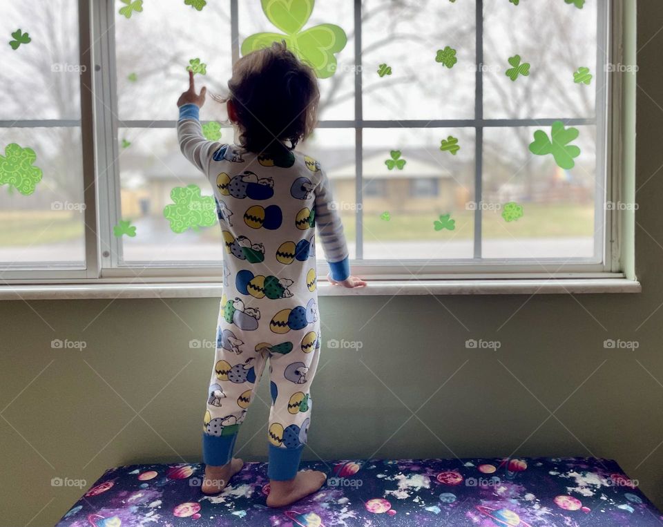 Toddler girl standing at the window on a bench pointing to shamrock decoration, toddler looking out window, toddler girl enjoys the shamrock decorations, decorating for the holidays, toddler girl watches over neighborhood, looking out of the window