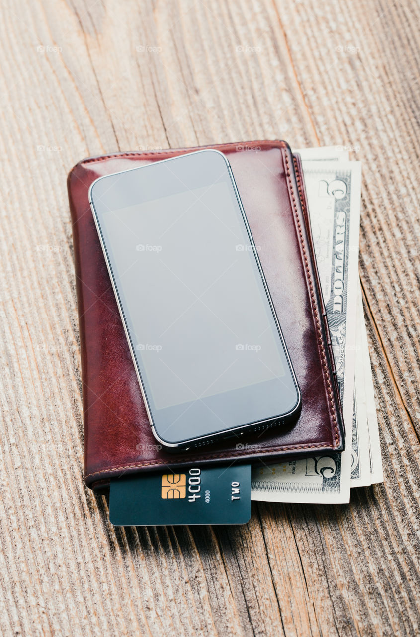 Smartphone with blank screen, wallet, dollar banknotes, debit credit cards and notebook on wooden table. View from above. Portrait orientation