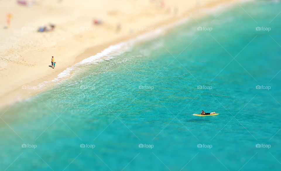 A view from above looking down onto a white sandy beach and emerald green ocean with small people and surf boarders enjoying the sunshine and summer vacation.