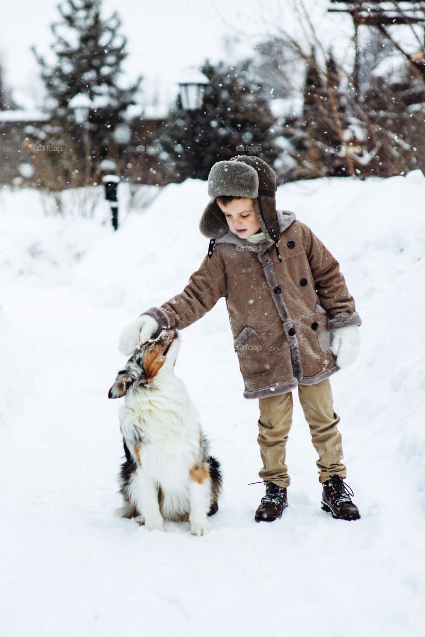 A boy in warm coat and winter hat strokes his dog. Australian Shepherd. Cute dog. A pet and its owner