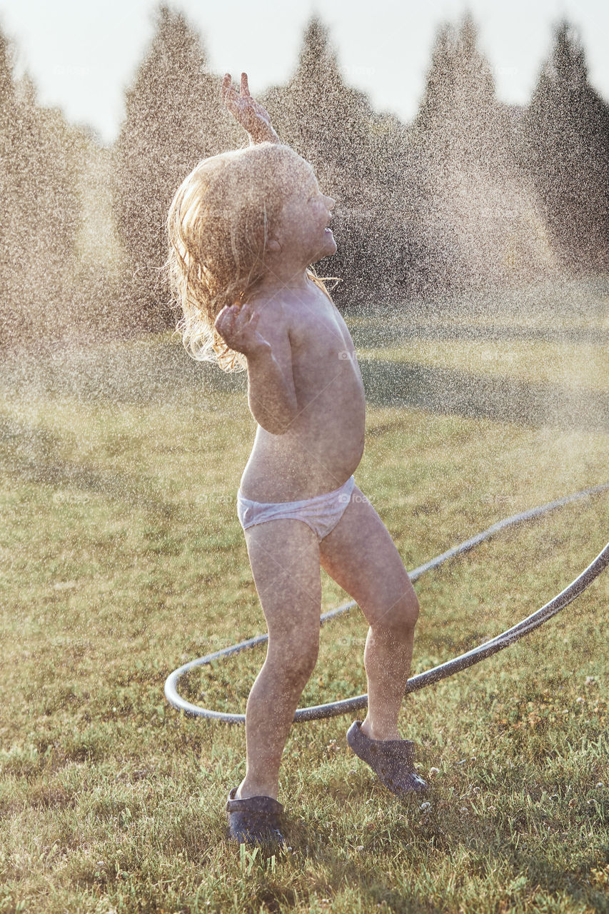 Little cute adorable girl enjoying a cool water sprayed by her mother during hot summer day in backyard. Candid people, real moments, authentic situations