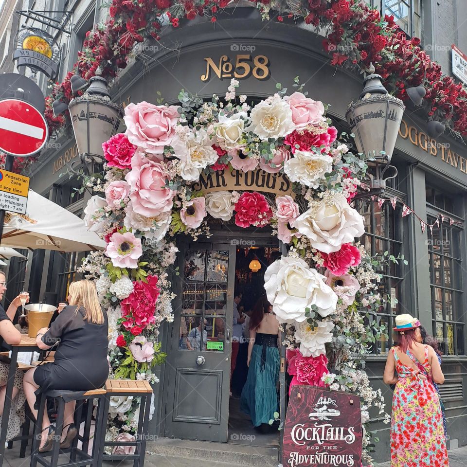 outside pub. floral display. summer day. summer dresses. people drinking outside in London. pink and white flowers around door