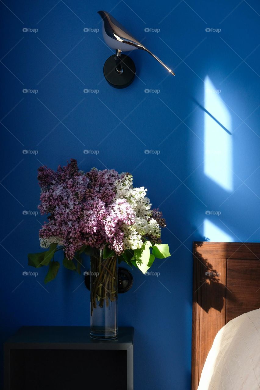 A bouquet of lilacs in the sunlight on a bedside table and golden bird shaped sconce in a blue bedroom