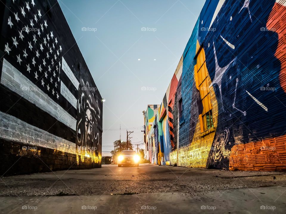 From the ground level, a car driving through an alley at dusk and it's headlights are illuminating street art on both left and right exterior walls.