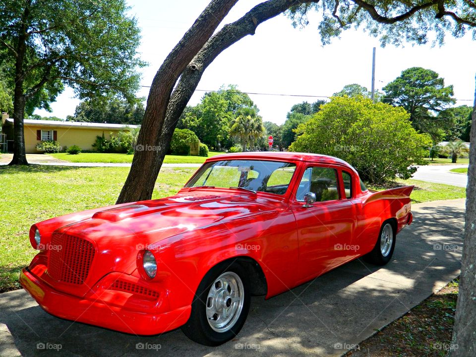 A beautiful red Golden Hawk Studebaker  is parked under a tree in the drive  