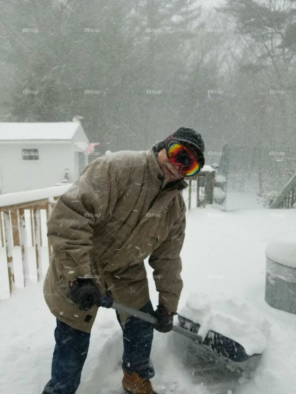 Dressed for snowstorm shoveling, skiing goggles, gloves, hat, shovel & smiling in the storm.