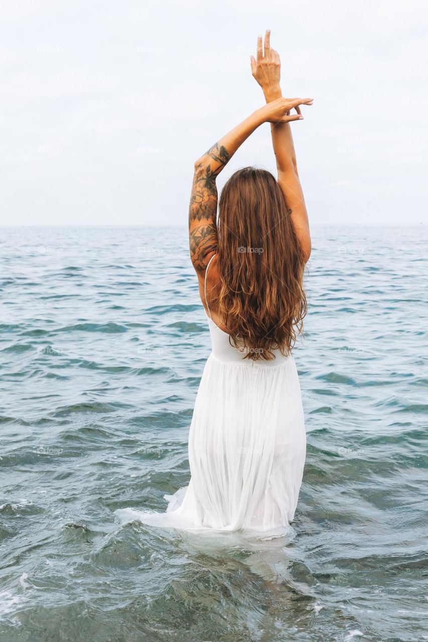 Young carefree woman with long hair in white dress enjoying life on sea beach, people from behind