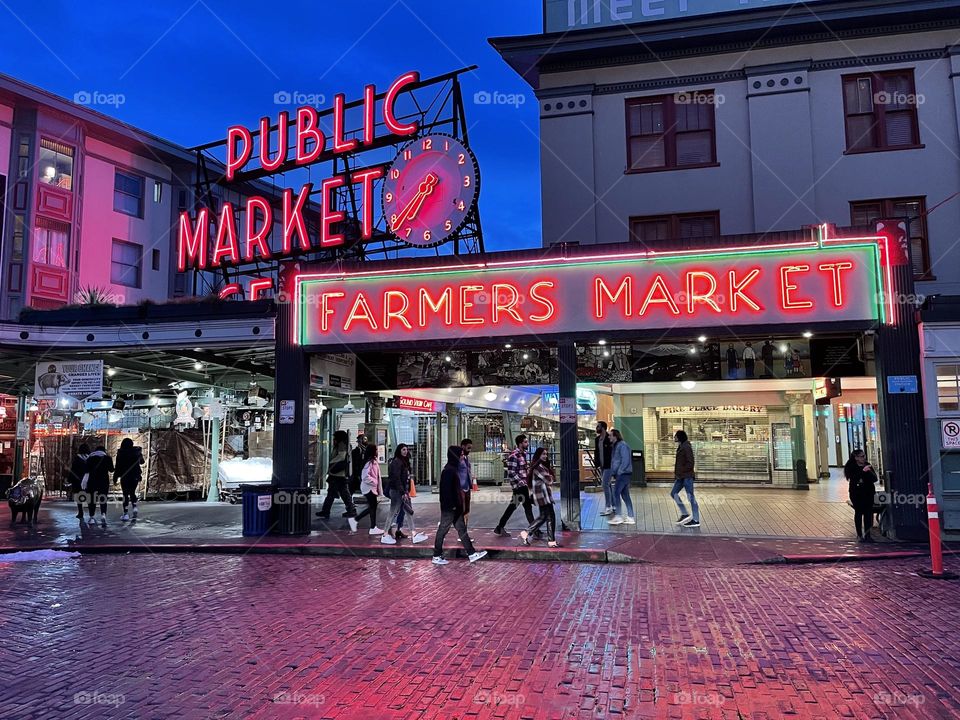 People milling about Seattle’s Public/Farmers Market at dusk. Neon signs reflect on wet cobblestone street
