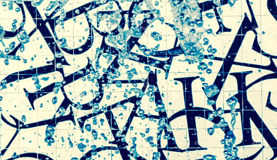 An off camera water fountain spray droplets in front of a tiled wall, which is patterned with a jumble of letters