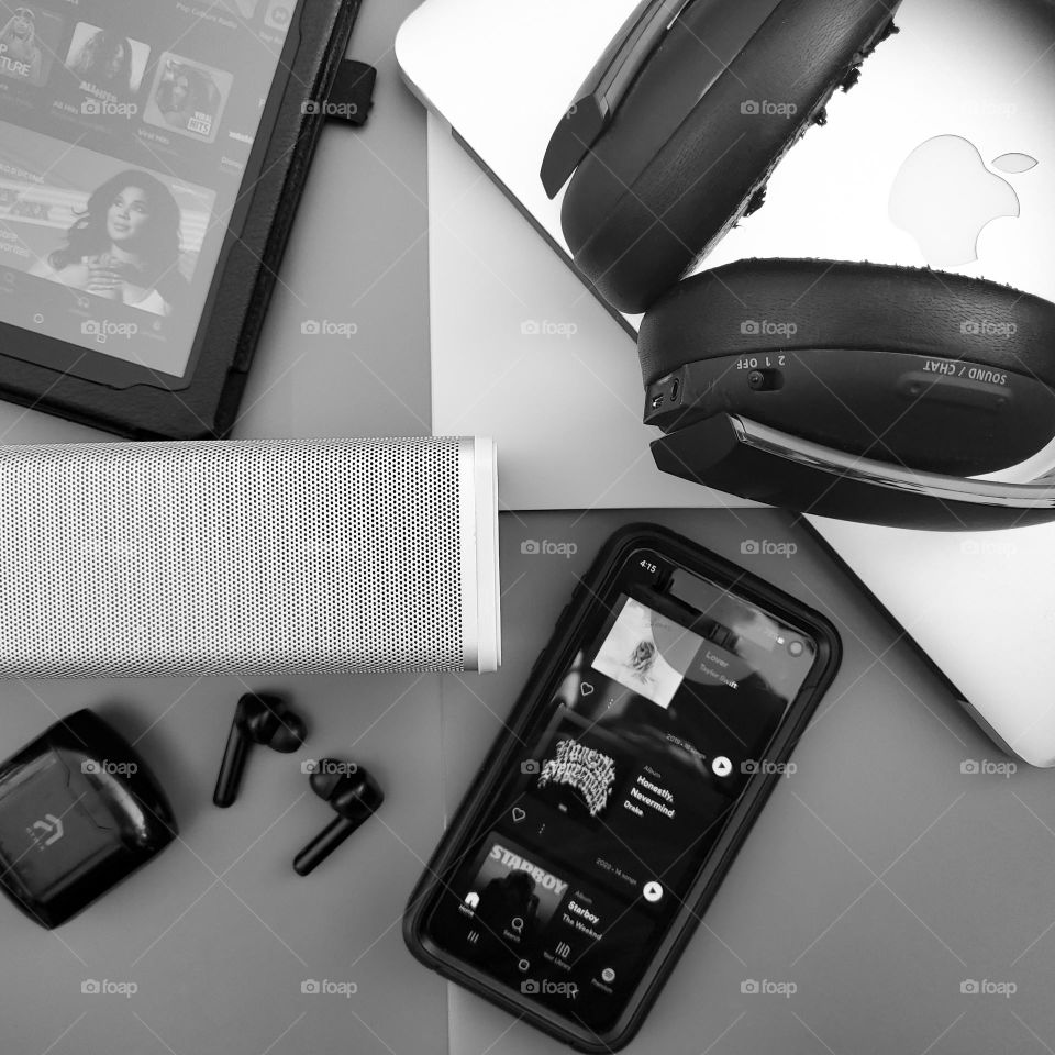 Electronic music listening devices black and white flatlay.  Tablet with Amazon music,  MacBook air,  Bluetooth headset,  Bluetooth ear buds, mobile phone,  and portable bluetooth speaker.