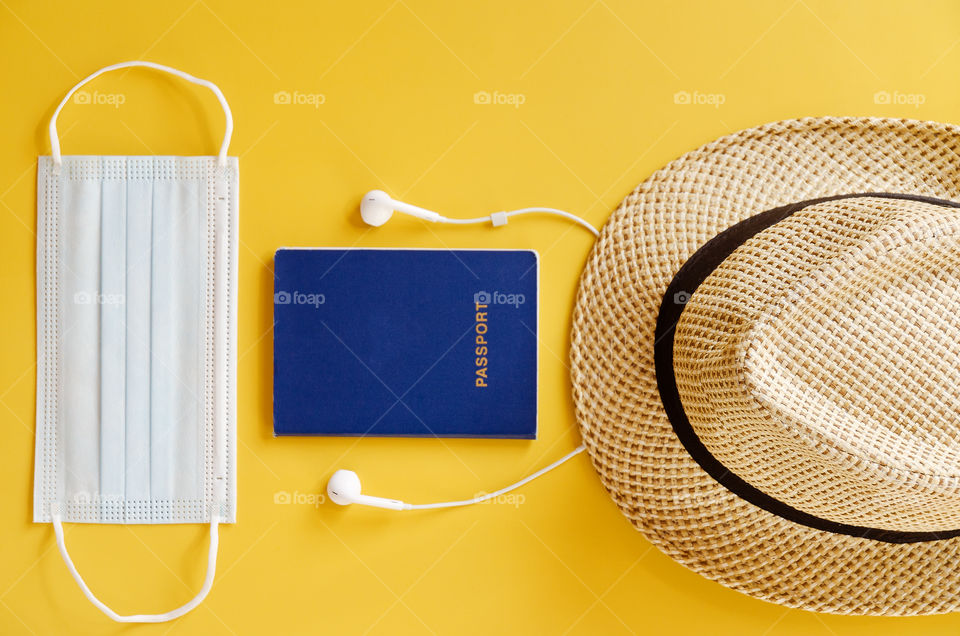 Travel after COVID-19 and new normal concept. Top view of medical mask, beach hat, passport, eyeglasses and headphones on yellow background close up. Travel concept.